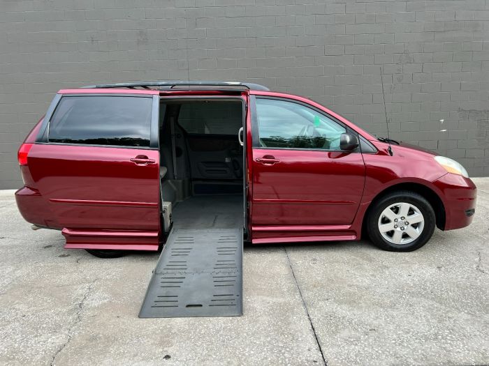 A red 2010 Toyota Sienna wheelchair van parked with the wheelchair ramp deployed from the passenger side sliding door. The backround is a gray block wall.