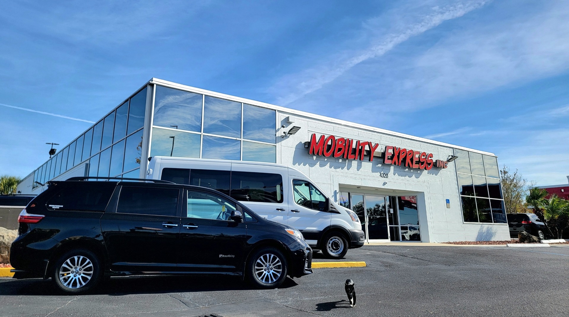 A white building with a lot of mirror windows. The words "Mobility Express" are on the front top of the building in red. There are two handicap vans, a dark colored minivan, and a white full size van, parked in front to the left.