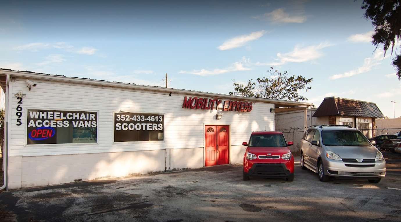 A white building with the words "Mobility Express" in red. There are two handicap accessible vehicles, a red small SUV and a silver minivan to the right.
