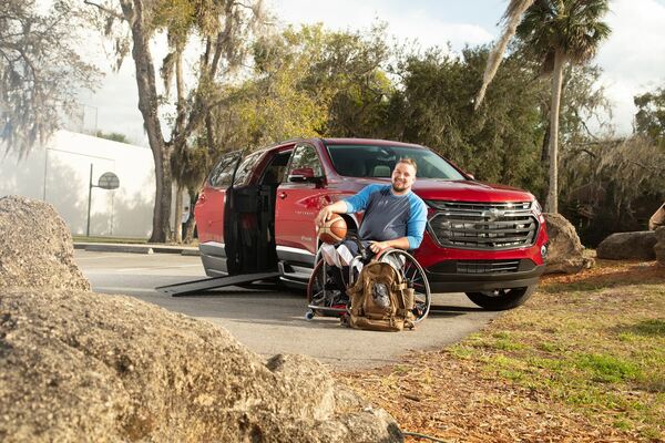Man in wheelchair in front of wheelchair accessible SUV with ramp open.