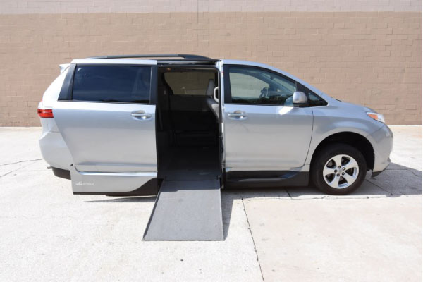 Silver Toyota Sienna wheelchair van with ramp deployed from open passenger side sliding door.